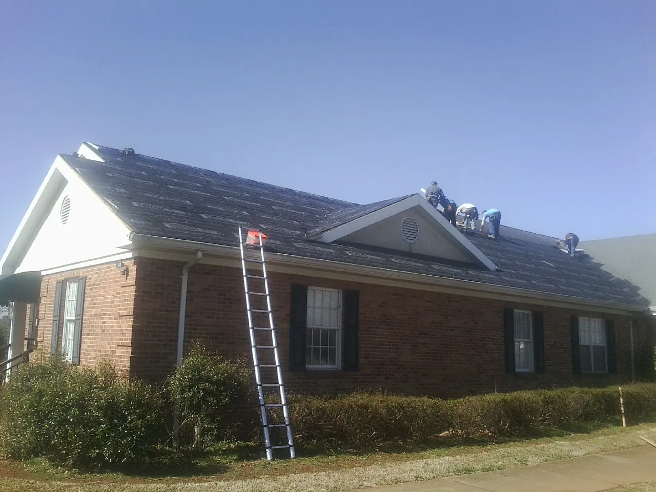 Crew tearing shingles off a brick ranch roof with a ladder set — Metal Roof Repair work by our crew in Macomb
